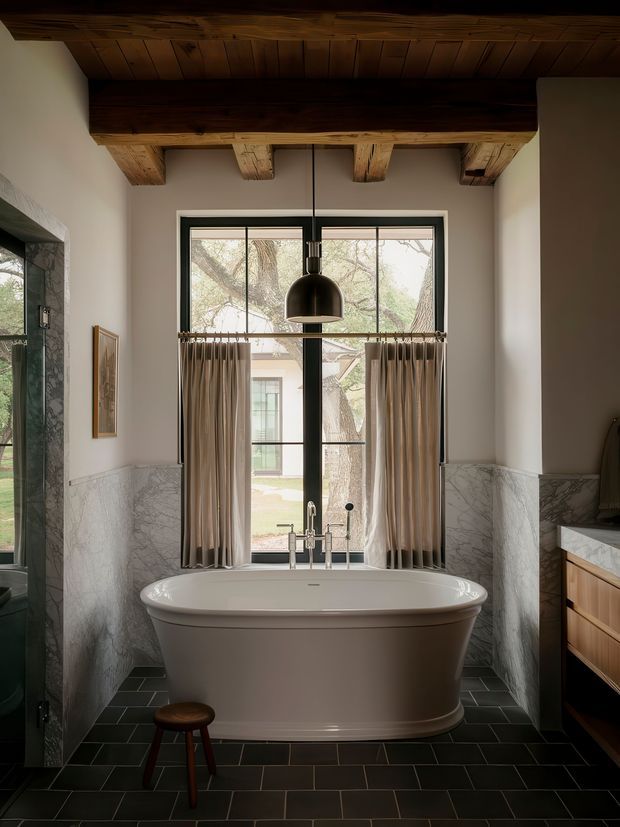 A sophisticated bathroom featuring a freestanding white bathtub positioned beneath a large black-framed window, with beige curtains and views to greenery beyond. The room has dark rectangular floor tiles, marble wall wainscoting, a timber vanity with a marble countertop, and a richly detailed exposed timber beam and plank ceiling. A black pendant light hangs centrally above the bath, creating a warm, refined atmosphere.