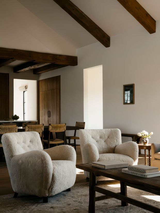Warm, refined open-plan living and dining area with white plaster walls, exposed dark timber ceiling beams, a wood dining table and chairs, two textured cream boucle armchairs, and a dark timber coffee table. Natural light enters through tall openings, creating a calm, earthy interior with traditional rustic architectural detailing and contemporary furnishings.
