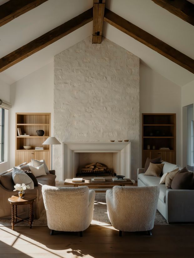 A warm, refined living room with a high vaulted ceiling, exposed dark timber beams, white plaster walls, and a large textured stone or plaster fireplace surround. Built-in timber shelving flanks the fireplace, while two sofas and two plush lounge chairs face a low wooden coffee table. Natural light enters from tall side windows, creating a cozy, elegant atmosphere.