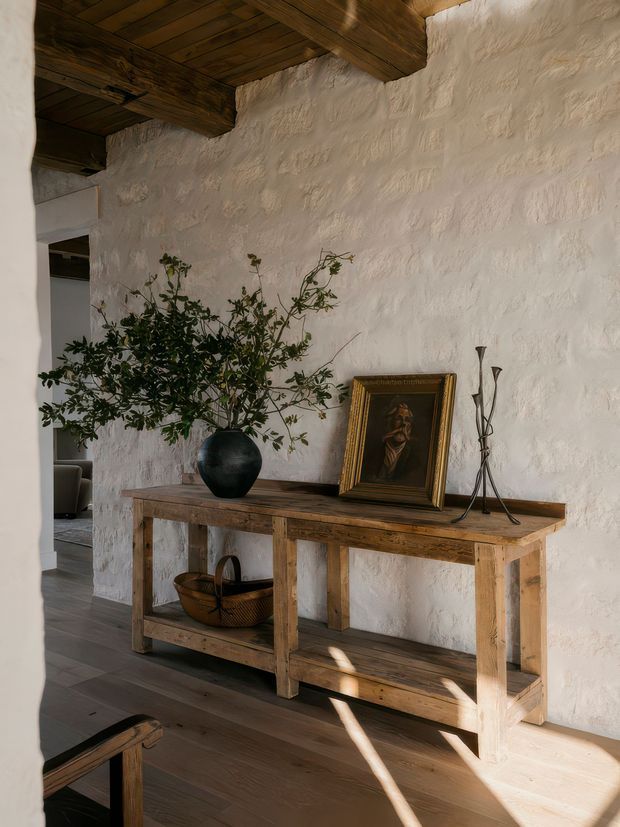 A warm, rustic interior vignette featuring a long reclaimed timber console table against a heavily textured white plaster wall. Exposed dark timber ceiling beams and planks frame the scene. The console is styled with a large black ceramic vase containing leafy branches, a framed portrait, a slender metal candle holder, and a woven basket on the lower shelf. An adjacent room is partially visible through an opening on the left.