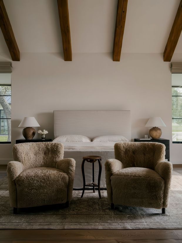 A warm, refined bedroom with a large upholstered bed, two black bedside tables, matching table lamps, and two plush boucle armchairs arranged in the foreground around a small dark wood side table. Exposed timber ceiling beams, pale walls, broad windows, layered neutral textiles, a soft area rug, and timber flooring create a calm, earthy interior.