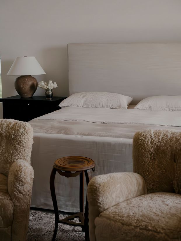 A warmly styled bedroom with a large upholstered bed, pale textured bedding, and a light neutral upholstered headboard. Two plush cream boucle armchairs frame a small dark wood side table in the foreground. A black bedside cabinet holds a sculptural ceramic lamp and a small floral arrangement, creating a soft, luxurious and layered interior.