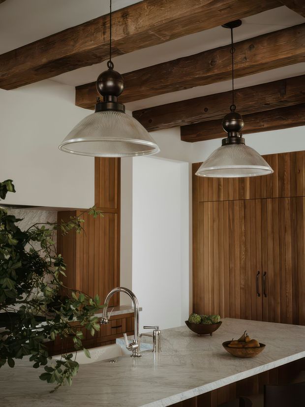 A warm, refined kitchen interior featuring a large pale stone island with an undermount sink and curved metal faucet, dark timber cabinetry with vertical panelling, exposed rough-hewn timber ceiling beams, and two pendant lights with ribbed glass shades and dark metal fittings. Greenery and bowls provide restrained natural accents.