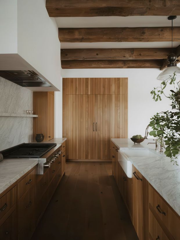 Warm galley kitchen with dark timber cabinetry, a matching timber floor and vertical timber pantry doors. White marble-look stone benchtops and splashback contrast with the wood, while exposed dark timber ceiling beams add rustic character. A large farmhouse-style sink, integrated cooktop and pendant lighting complete the space.