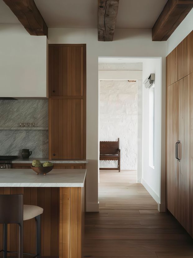 A warm, refined kitchen interior viewed toward a bright hallway and adjoining room. The kitchen features vertical dark timber cabinetry, a pale stone island benchtop, grey veined stone splashback, timber flooring, and exposed dark timber ceiling beams. Full-height fluted timber cabinetry lines the right wall, while a bright secondary room is visible through the doorway.