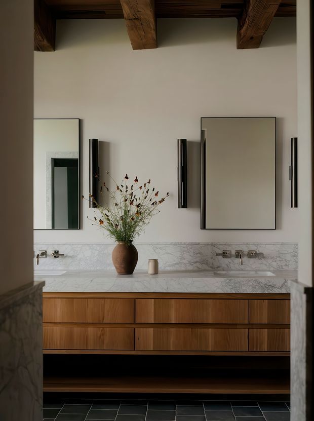A refined bathroom vanity viewed through a doorway, featuring a long double-sink timber vanity with fluted drawer fronts, a white marble benchtop and backsplash, wall-mounted brushed-metal tapware, two large rectangular mirrors, slim vertical sconces, and a dark tiled floor. Exposed rustic timber ceiling beams add warmth and character.