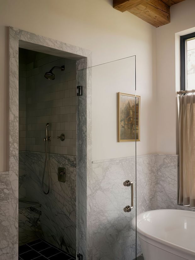 Elegant bathroom interior featuring a marble-lined walk-in shower with dark tiled flooring, exposed timber ceiling beams, marble wainscoting, a frameless glass shower screen, and a freestanding white bathtub beside a curtained window. Warm natural light and traditional detailing create a refined, luxurious atmosphere.