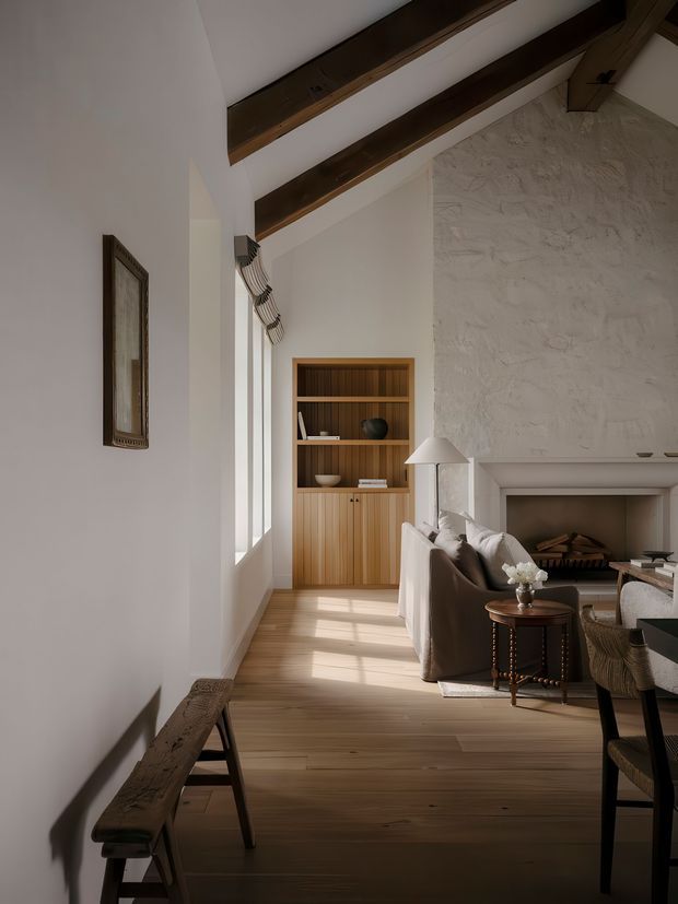 A refined living room with a high pitched ceiling, exposed dark timber beams, white plaster walls, a textured masonry fireplace, built-in timber shelving and cabinetry, and wide timber flooring. Natural light enters through tall windows, illuminating a neutral seating area and small side table.