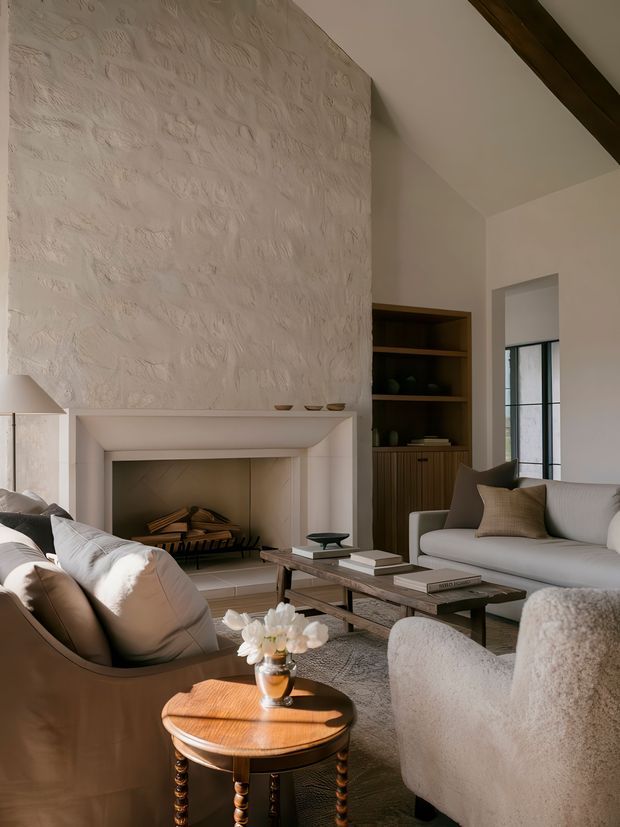 A warm, refined living room centred around a large white sculptural plaster fireplace with a textured feature wall. The room includes a pale sofa, curved upholstered lounge chairs, a dark wood coffee table, a round timber side table, built-in dark wood shelving, and exposed dark ceiling beams. Soft natural light enters through a black-framed window, creating a calm, layered interior.