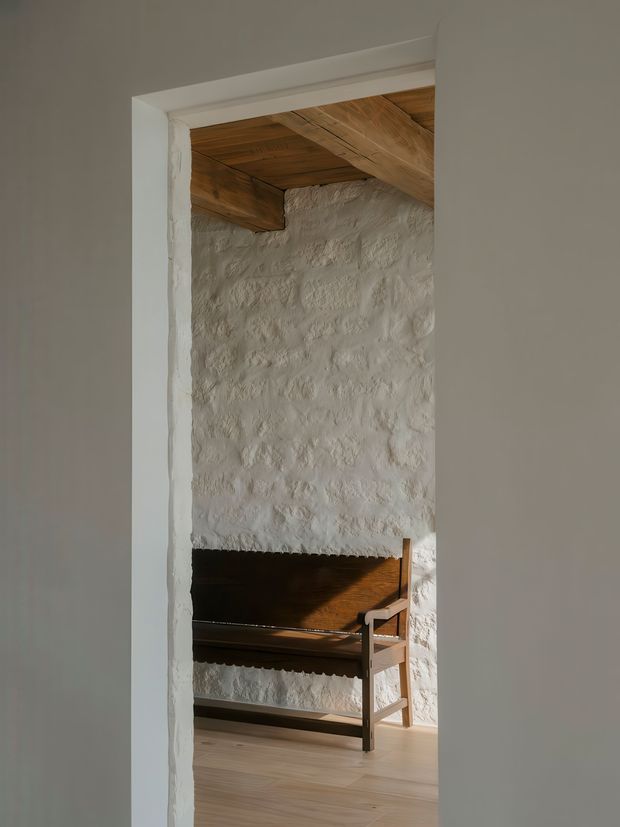 A narrow interior view through a white-painted doorway into a rustic room. The space features a rough white masonry wall, exposed timber ceiling beams and timber ceiling boards, pale timber flooring, and a dark wooden built-in or freestanding bench with a simple ladder-like side structure. Warm sunlight creates strong shadows across the wall and furniture.