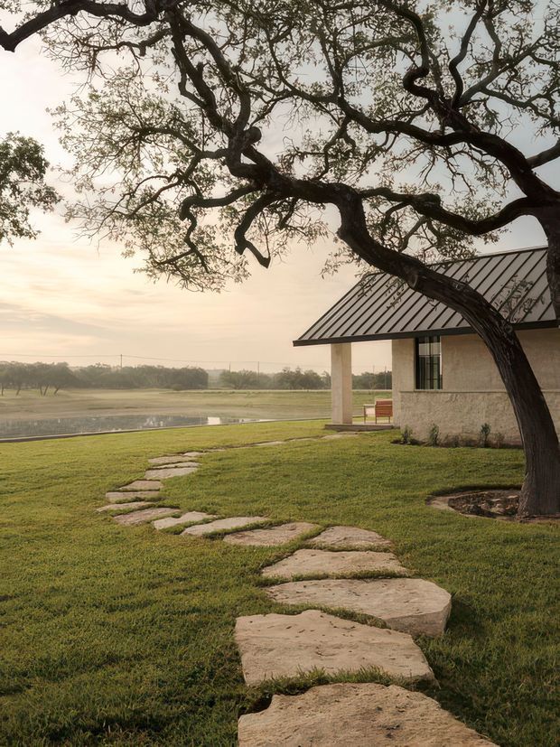 A tranquil rural exterior view of a low, single-storey building beside an open grassy landscape. A large mature spreading tree frames the scene, while an irregular stepping-stone path leads across the lawn toward the building. The structure has a simple pale masonry facade, dark standing-seam pitched roof, covered porch supported by square columns, and tall narrow glazing. A reflective water feature or pond appears beyond the lawn.
