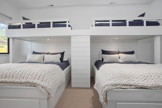 Bright, compact bedroom featuring two built-in lower beds with white textured bedding, navy and white pillows, and two elevated bunk-style sleeping platforms above. White horizontal wall panelling and built-in cabinetry create a clean, highly integrated design, with neutral carpet and small high windows.