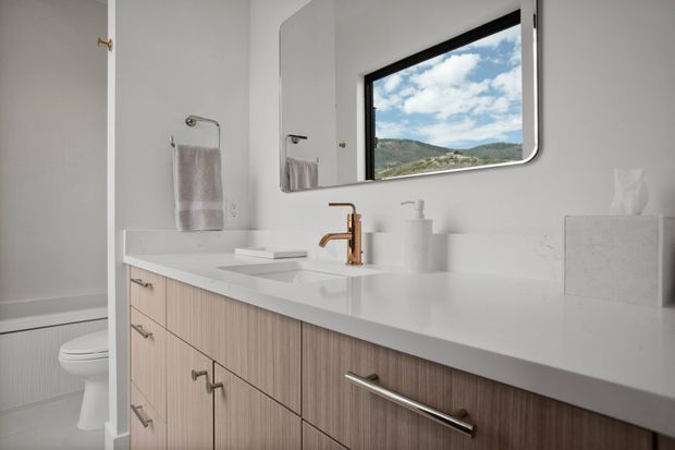 Bright contemporary bathroom with a long white vanity countertop, vertically grained light-wood cabinetry, brushed metal hardware, and a warm brass faucet. A toilet, towel rails, soap dispenser, and tissue box are visible, with a large rounded-corner window framing a mountainous landscape.