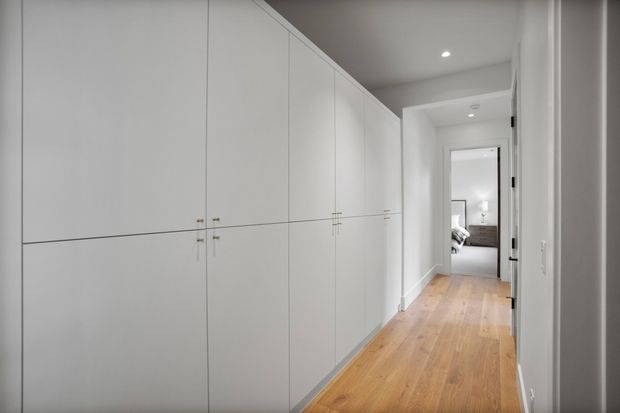 A bright, minimalist hallway with extensive full-height white built-in cabinetry along the left wall, warm timber flooring, white walls and recessed ceiling lights. An open doorway at the end provides a glimpse into a furnished bedroom or living space.