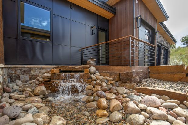 Exterior view of a contemporary building with dark cladding, warm timber soffits and wall panels, and a metal-railed entry terrace above a landscaped dry creek bed with rounded river stones and a small cascading water feature.