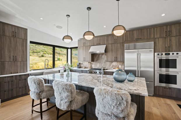 A spacious contemporary kitchen with a large marble-patterned island, dark wood cabinetry, integrated stainless-steel appliances, and three pendant lights. Floor-to-ceiling corner glazing brings in abundant natural light and overlooks a dry, hilly landscape. The island is paired with three plush furry bar stools, while warm timber flooring and neutral finishes create a refined, inviting atmosphere.