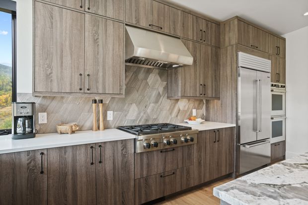 A contemporary kitchen with extensive wood-grain cabinetry, a white countertop, stainless-steel range and hood, integrated double wall ovens, and a large stainless-steel refrigerator. The backsplash uses a light grey geometric herringbone tile pattern, while a marble-look island occupies the foreground. A window provides a scenic view beyond the kitchen.