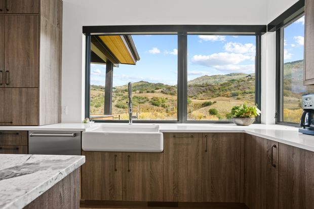 Bright contemporary kitchen with warm wood cabinetry, a white farmhouse-style sink, pale stone countertops, and large black-framed windows overlooking a dry rolling hillside landscape. A partial island with a marble-patterned surface and integrated appliances are visible.