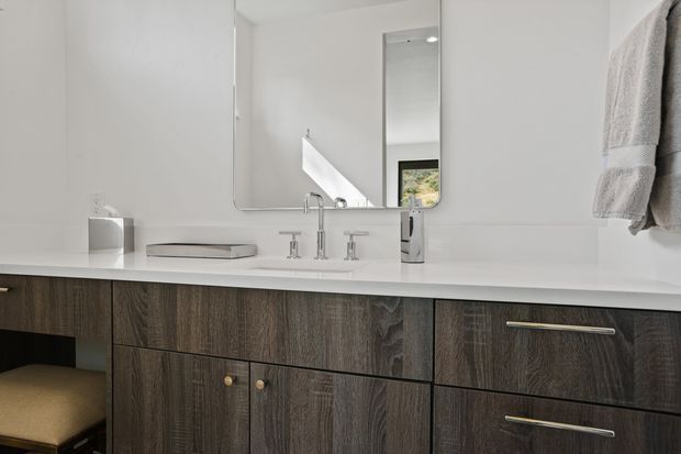 Bright contemporary bathroom vanity with a long white benchtop, dark wood-grain cabinetry, wall-mounted mirrored cabinet, twin basins and chrome tapware. A towel hangs on the right, and a stool is partially visible beneath the vanity.