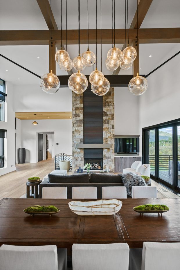 A bright, spacious open-plan living and dining area with a dramatic double-height ceiling. Exposed dark timber beams support a cluster of clear glass pendant lights above a large dark timber dining table. The living room includes a sectional sofa, television, and tall stone-clad fireplace with a dark metal feature insert. Black-framed floor-to-ceiling glazing opens toward a balcony and scenic landscape.