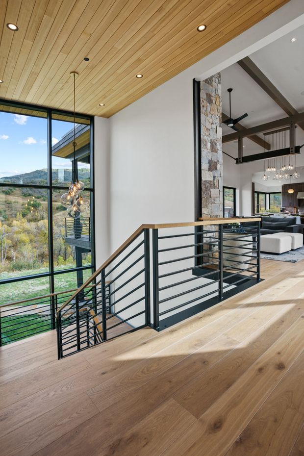 A bright, open-plan upper-level landing overlooking a stairwell, with wide-plank timber flooring and a matching timber-lined ceiling. Black metal horizontal railings border the stairs, while floor-to-ceiling glazing provides expansive views of a wooded hillside. A stone-clad structural column, exposed dark beams, contemporary pendant lighting, and part of a living room are visible beyond.