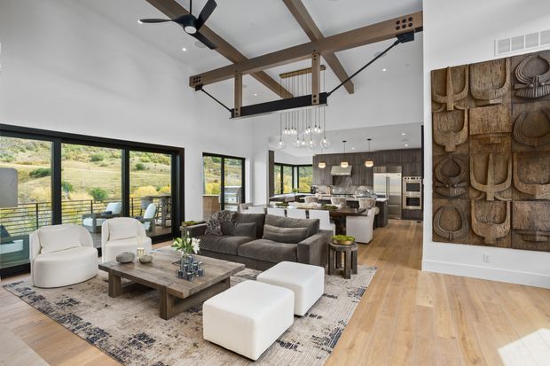 Open-plan contemporary living room with a high exposed-beam ceiling, large black-framed windows, and views to a landscaped hillside. The room includes a dark sectional sofa, white lounge chairs and ottomans, a timber coffee table, patterned area rug, sculptural wall art, and a kitchen with island beyond.