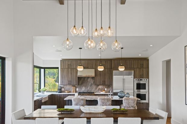 Bright open-plan kitchen and dining area with extensive dark wood cabinetry, a stone island and backsplash, stainless-steel appliances, and a large dark timber dining table. Globe pendant lights hang from a white ceiling, while upholstered bar stools and dining chairs add a soft contemporary feel. Large windows bring in natural light and views of greenery.