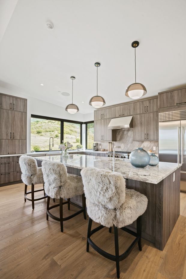 Bright, spacious contemporary kitchen with a large marble-look island and three upholstered bar stools. The kitchen features extensive wood-grain cabinetry, integrated stainless-steel appliances, a sculptural rangehood, black-framed corner glazing, timber flooring, and three pendant lights. A landscaped hillside is visible through the windows.