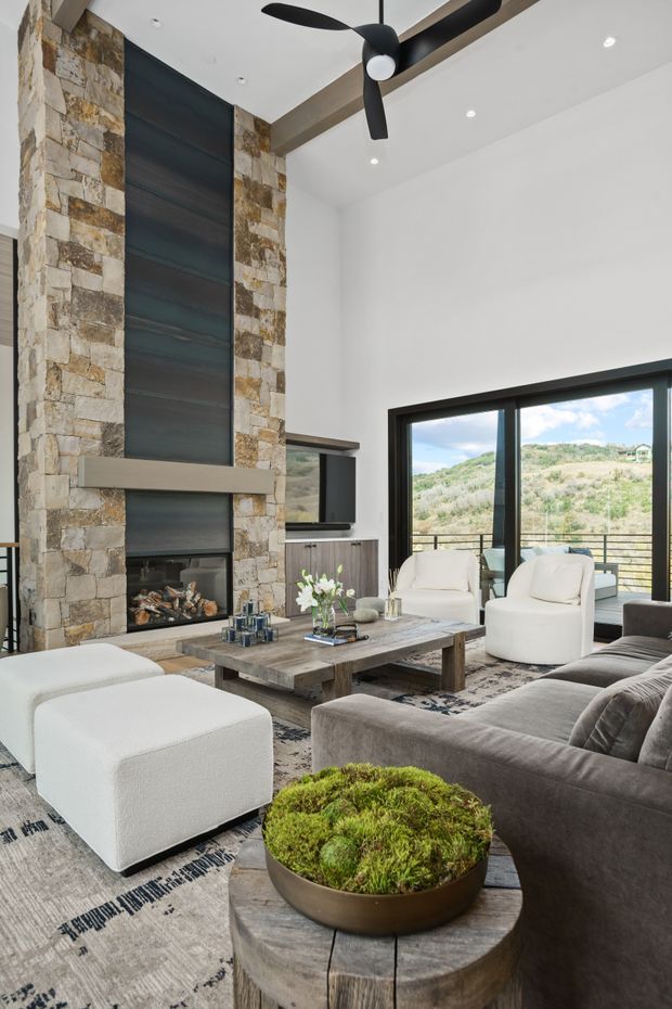 A contemporary living room with a tall stacked-stone fireplace wall, dark metal fireplace surround, exposed timber ceiling beam, black ceiling fan, and expansive black-framed glazing opening to a scenic hillside view. The room is furnished with a large grey sectional sofa, white upholstered lounge chairs and ottoman, a rustic timber coffee table, patterned rug, and a round timber side table with a moss planter.