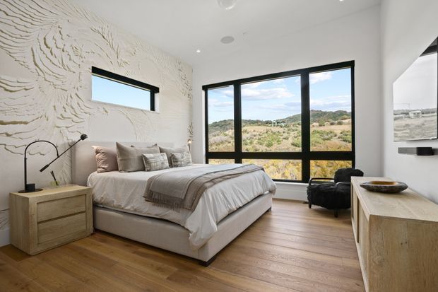 Bright contemporary bedroom with a large upholstered bed, layered neutral bedding, pale timber flooring, sculptural textured wall finish, black-framed windows, and a light timber bedside table and dresser. The expansive glazing overlooks a dry, landscaped natural setting.