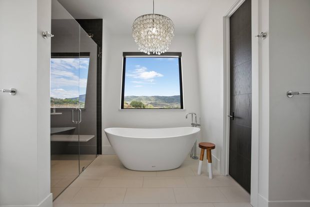 Bright contemporary bathroom with a freestanding white oval bathtub beneath a decorative pendant light, large window overlooking rolling hills, glass-enclosed shower with dark tiled walls, pale floor tiles, and a dark timber door.
