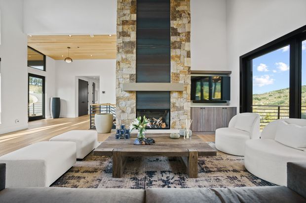Bright, spacious contemporary living room with a large stone-clad fireplace, timber mantel and hearth, white upholstered seating, a substantial rustic timber coffee table, and expansive black-framed glazing overlooking a green hillside. The room includes a timber-lined ceiling zone, dark wood cabinetry, and a patterned area rug, creating a refined natural-materials interior with strong indoor-outdoor connection.