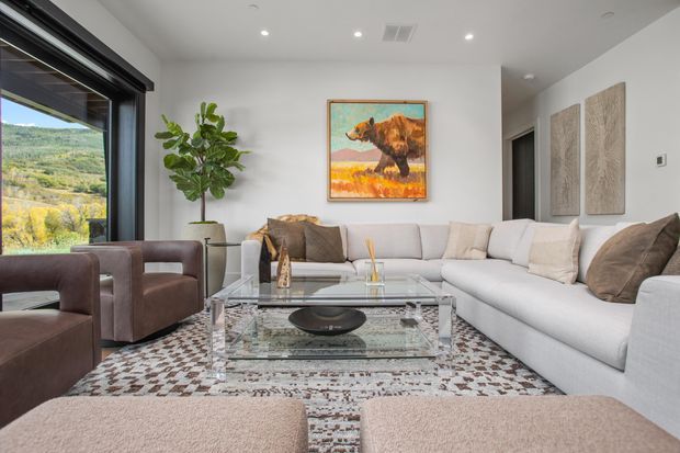 A bright, contemporary living room with a large white sectional sofa, brown upholstered armchairs, a transparent glass coffee table, patterned area rug, and neutral upholstered ottomans. Floor-to-ceiling black-framed glazing opens to a scenic landscape, while a bold animal painting and decorative wall panels add visual interest.