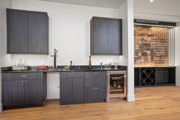 Contemporary open-plan kitchen and home bar area with dark vertically textured cabinetry, dark stone benchtops, integrated sink and cooktop, and a stainless-steel under-bench wine refrigerator. Warm timber flooring contrasts with white walls. To the right, a built-in wine storage and display area features a black diamond wine rack and a decorative tiled or gridded backsplash.