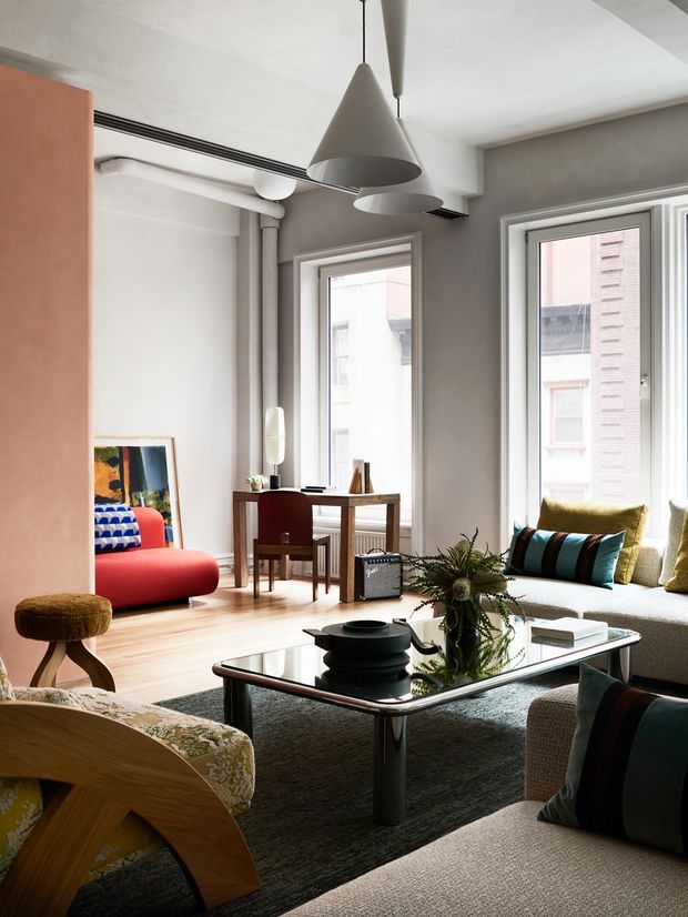 A bright, contemporary living room in an apartment-like setting, featuring tall windows, pale timber flooring, grey walls, exposed service pipes, and sculptural pendant lights. The room contains a low glass coffee table, upholstered sofas, a curved wooden lounge chair, a red accent chair, a compact writing desk, and colourful cushions. A large indoor plant and artwork add visual interest, while neighbouring buildings are visible through the windows.
