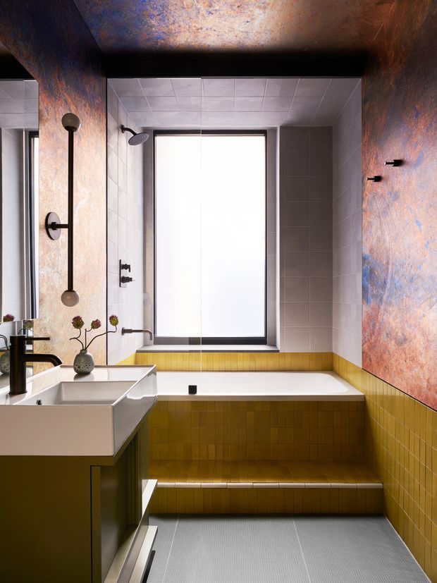 A compact, highly designed bathroom with a built-in soaking tub set into a stepped mustard-yellow tiled platform. The room features pale grey wall tiles, a large frosted window with dark framing, a sculptural white vessel sink on dark cabinetry, black plumbing fixtures, and dramatic mottled blue, copper, and rust-toned feature surfaces on the ceiling and side walls. The composition is bold and graphic with strong colour and material contrast.