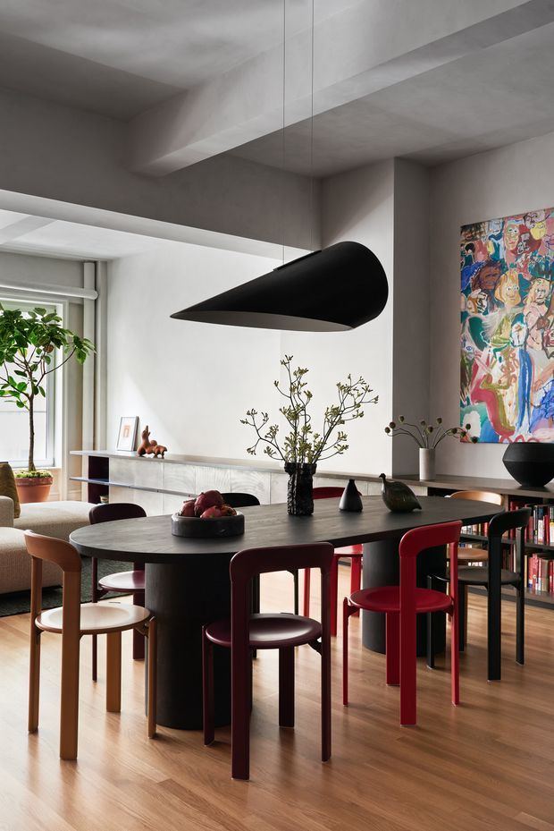 A refined contemporary dining area with a large dark oval dining table surrounded by sculptural chairs in burgundy, black and timber tones. A dramatic black pendant light hangs over the table, while built-in shelving, a low sideboard, indoor plants, ceramics and colourful artwork add character. The room has warm timber flooring, pale textured plaster walls and a partially visible adjoining living area with generous glazing.