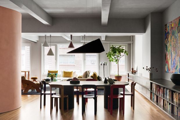 A bright open-plan contemporary dining and living space with a large dining table surrounded by mismatched colourful chairs, a lounge area beyond, extensive windows, a sculptural black pendant light, exposed concrete ceiling beams, timber flooring, and a long built-in media and bookcase wall. A curved pink partition and colourful wall artwork add visual interest.