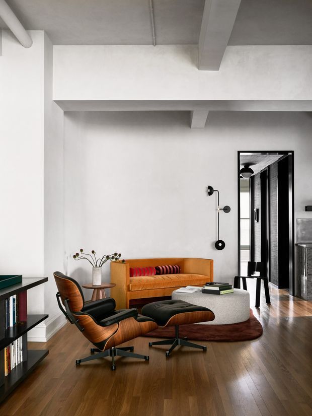 A refined living room with a high exposed concrete ceiling and structural beams, white plaster walls, dark timber flooring, and black-framed glazing or doors. The room is furnished with a warm tan sofa, sculptural mid-century lounge chair and ottoman, a rounded light upholstered coffee table, a dark red rug, and minimalist black shelving. A graphic wall sconce and colourful cushions add restrained accents. The open doorway leads to another interior area.