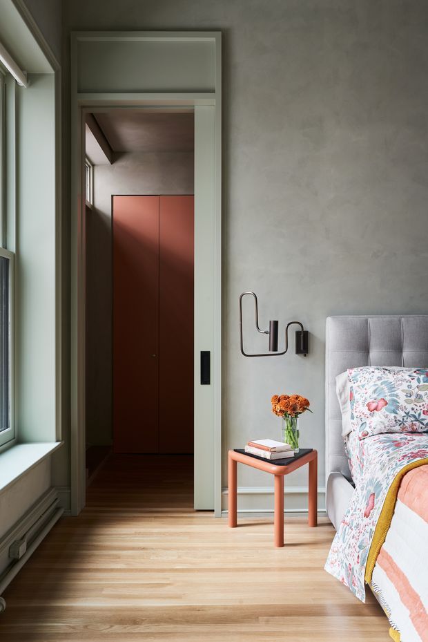 A refined bedroom interior with a grey upholstered bed, colourful floral bedding, pale grey textured walls, and warm timber flooring. A small coral-toned bedside table with flowers and books sits beside the bed, while a partially open doorway reveals dark reddish-brown built-in cabinetry in the adjacent space. A large window brings in natural light, and sculptural black wall lighting adds a contemporary accent.