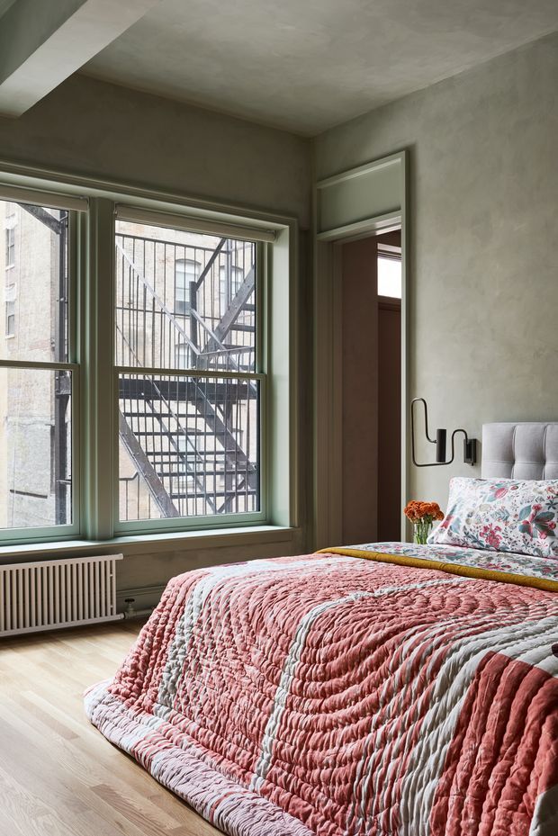 A softly lit bedroom with a large upholstered bed dressed in a textured pink, white and grey quilt and floral cushions. The room has pale timber flooring, muted green-painted window and door frames, a white radiator, plastered walls and ceiling, and a simple black wall-mounted reading light. Large industrial-style windows overlook an external fire escape, giving the room an urban character.