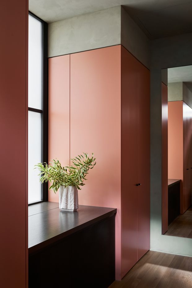 A refined interior joinery and wardrobe area with tall muted terracotta-pink cabinetry, a dark timber or stained-wood built-in bench beneath a narrow black-framed window, and a full-height mirror reflecting the cabinetry. The space has a smooth concrete-look ceiling and upper wall surfaces, dark window framing, warm timber flooring, and soft natural light. A leafy plant in a textured vase provides a small organic accent.