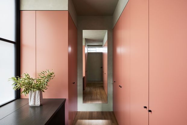 A narrow, refined residential hallway lined with full-height salmon-pink built-in cabinetry. A large full-height mirror at the end reflects the corridor and timber flooring, while a dark timber console beneath a tall black-framed window holds a potted plant. The space has a warm, muted palette and clean contemporary detailing.