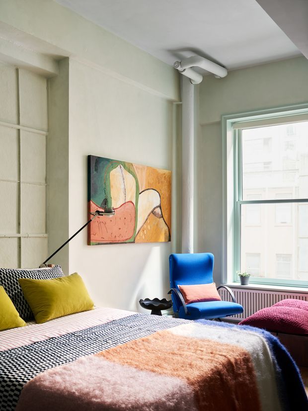 A bright, eclectic bedroom with pale green walls, a large window, exposed white-painted pipes, and a layered bed dressed in patterned black-and-white bedding, mustard and pink cushions, and a soft peach throw. A vivid blue armchair with a pink cushion sits beside the window, with colourful abstract artwork above the bed and a small sculptural side table. The room has a playful, art-led interior with strong contrasting colours and abundant natural light.