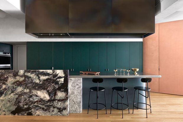 A dramatic contemporary kitchen with a long island and three black bar stools. The island features a highly patterned dark natural-stone waterfall end and a pale stone or solid-surface benchtop. Behind it is a wall of deep teal-green flat-panel cabinetry, partly concealed appliances, and a large dark overhead rangehood enclosure. A salmon-pink wall and light timber flooring add contrast, while black-framed fixtures and stools create a refined, graphic look.
