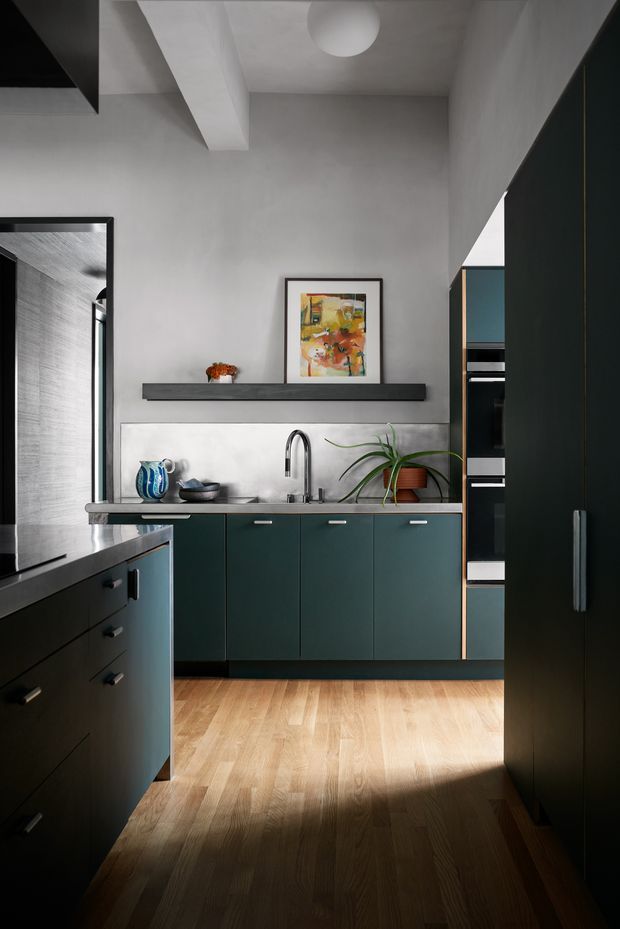 A contemporary galley-style kitchen viewed from the entry, featuring dark teal-green lower cabinetry, tall dark cabinetry with integrated ovens, a pale stone or concrete-look splashback, and a dark benchtop. Warm timber flooring contrasts with the predominantly charcoal, teal, and light grey palette. A sculptural pendant light, artwork, open shelving, a potted plant, and ceramic vessels add character. The room has a high ceiling with exposed-looking beams and strong natural light creating a dramatic foreground shadow.