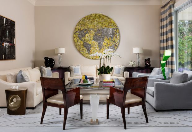 A bright, refined living room arranged around a glass coffee table, with cream sofas and armchairs, dark wood dining-style chairs, a grey lounge chair, table lamps, artwork, an orchid, and a large floor-to-ceiling window with striped drapery. The space has a geometric light-toned floor and a curated mix of traditional and modern furnishings.