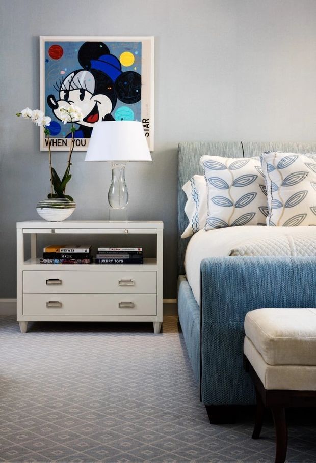 A styled bedroom corner with a blue upholstered bed, patterned blue-and-white bedding and cushions, a white bedside cabinet with drawers and open shelving, a glass table lamp, and an orchid in a striped planter. A colourful framed artwork hangs above the cabinet. The room has a soft grey wall and patterned grey carpet, creating a polished, comfortable interior.