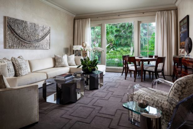 Elegant, traditionally detailed living room with a dining area by tall multi-pane windows overlooking lush greenery. The room features a cream sectional sofa, patterned cushions, dark geometric coffee tables, a patterned area rug, sculptural wall art, and a glass-and-patterned accent chair. Decorative ceiling mouldings, full-height curtains, and refined furnishings create a luxurious, polished atmosphere.