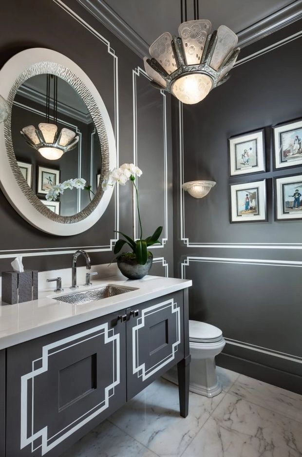 A dramatic, highly decorated powder room with charcoal-grey walls and vanity cabinetry outlined in crisp white geometric moulding. The room features a white stone countertop with an undermount basin, polished metal tapware, a large oval textured mirror, ornate sculptural wall and ceiling lights, framed artwork, and a white toilet. Veined light stone flooring and a potted orchid add contrast. The overall look is glamorous and Art Deco-inspired.